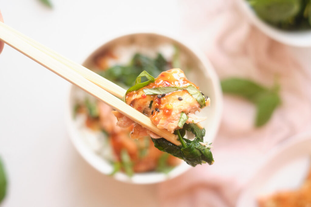 Chopsticks holding sesame salmon and spinach and basil above an Asian rice bowl