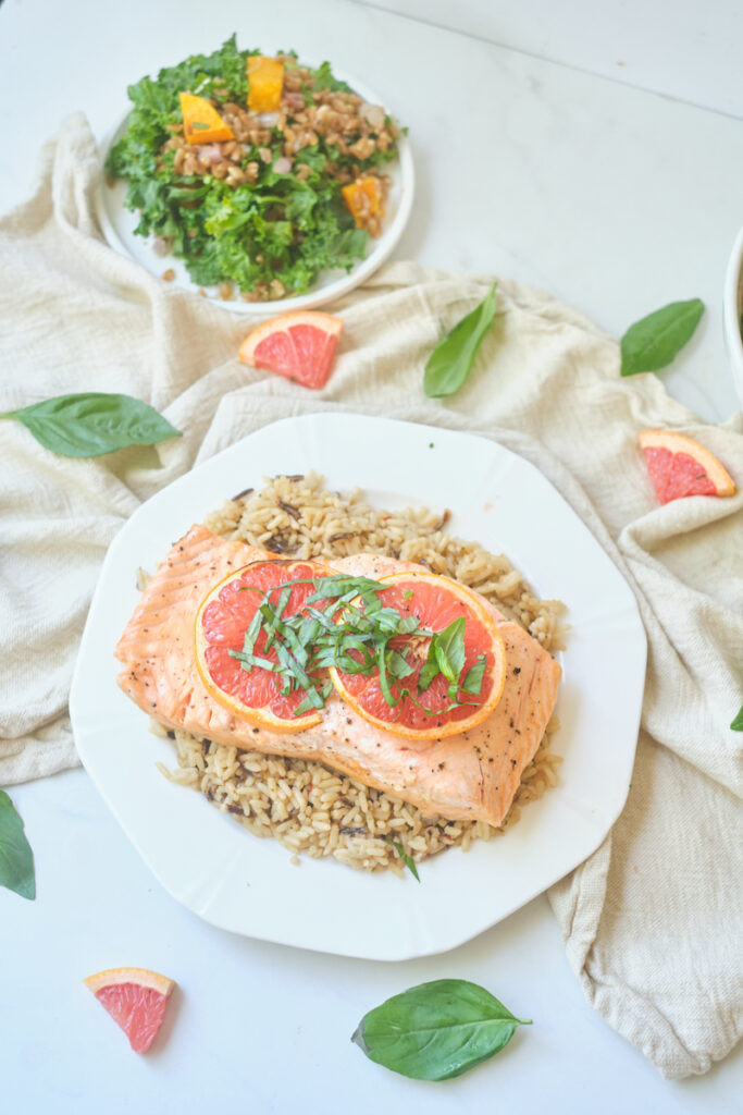 baked grapefruit salmon with grapefruit slices and basil chiffonade on a plate over brown rice, with a hearty salad in the background