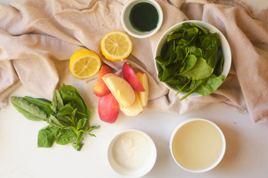 Ingredients for an apple basil matcha protein smoothie on a table including spinach, apple, lemon, basil, matcha, protein powder and plain greek yoghurt