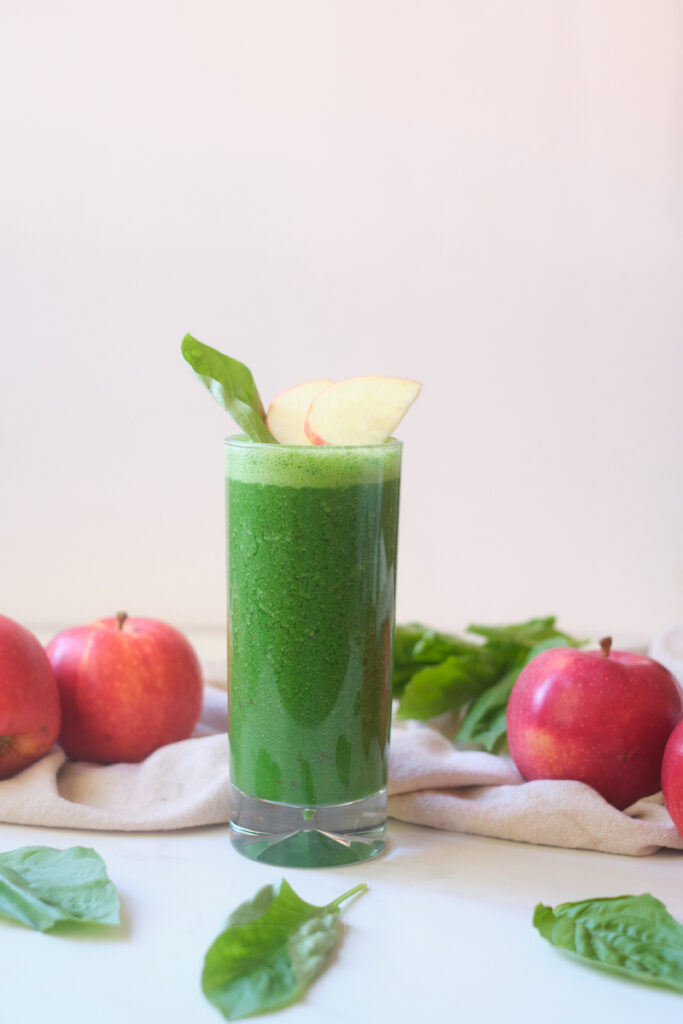 An apple basil matcha protein smoothie in a glass, with a basil leaf and apple wedges sticking out the top
