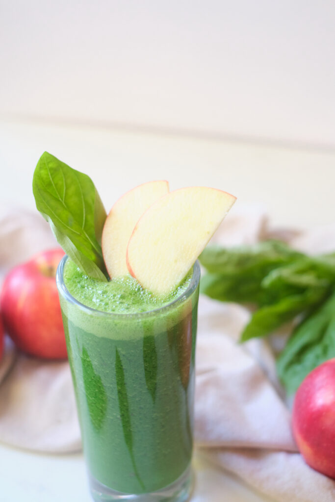 An apple basil matcha protein smoothie in a glass, with a basil leaf and apple wedges sticking out the top