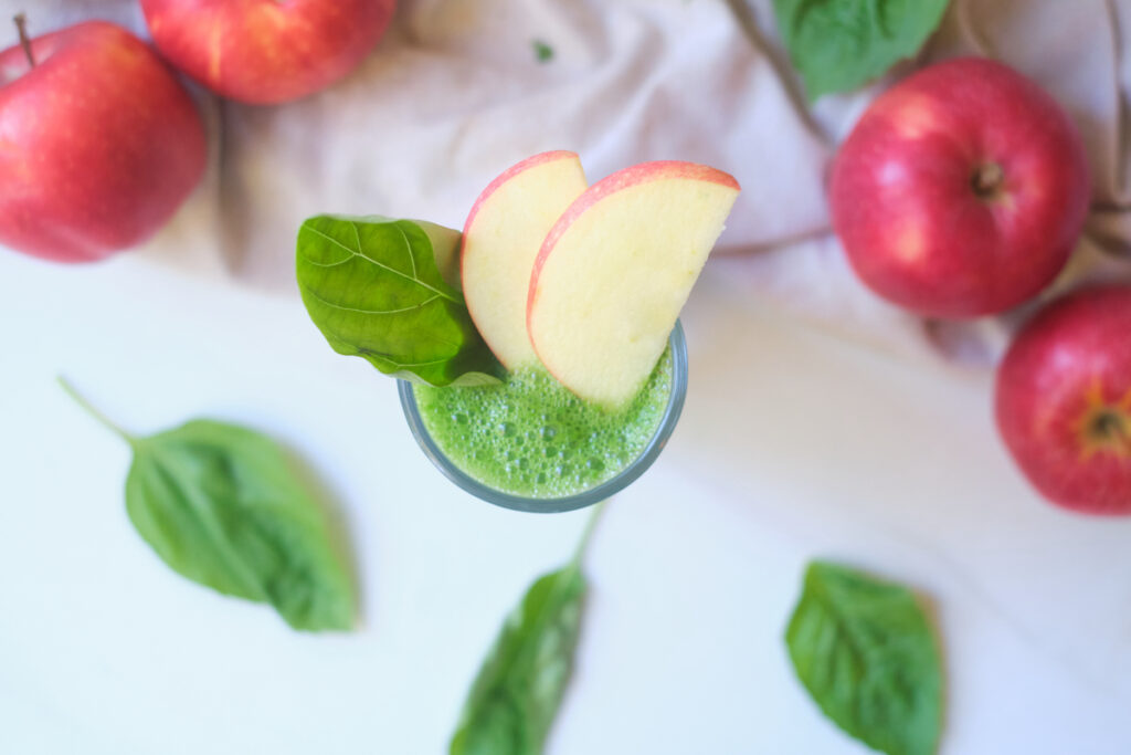 birds eye view of an apple basil green smoothie in a glass, with apples and basil leaves strewn about around the glass