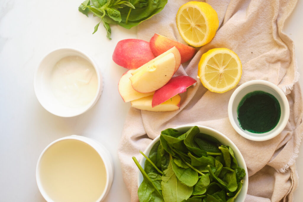 Ingredients for an apple basil matcha protein smoothie on a table including spinach, apple, lemon, basil, matcha, protein powder and plain greek yoghurt