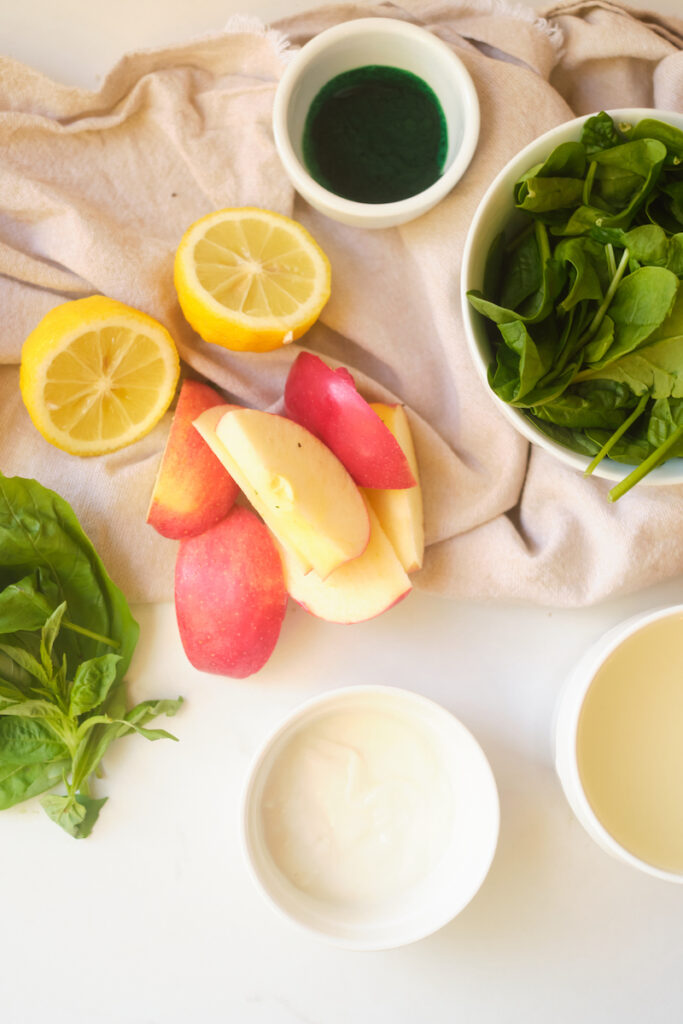 Ingredients for an apple basil matcha protein smoothie on a table including spinach, apple, lemon, basil, matcha, protein powder and plain greek yoghurt