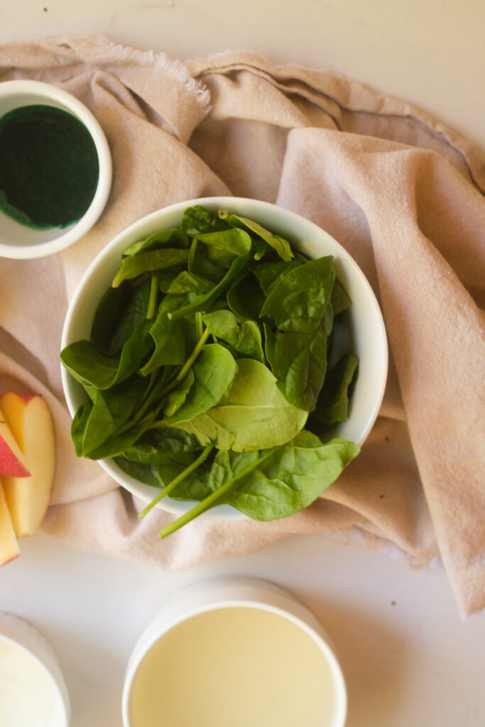 Close up of a bowl of spinach with some other ingredients for an apple basil green smoothie in view