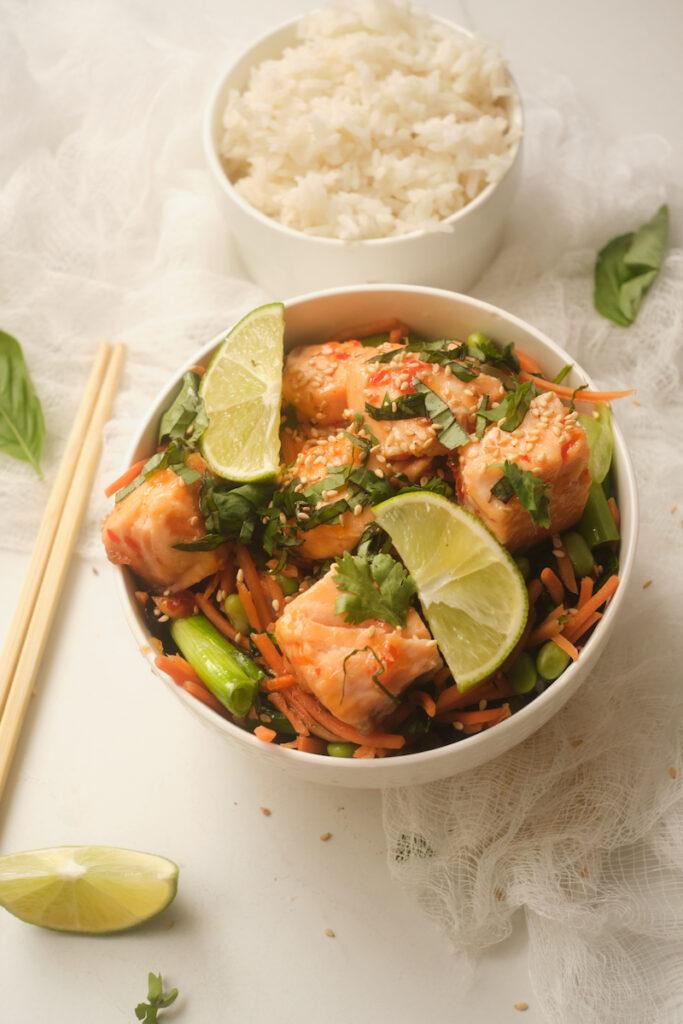 Sweet chilli salmon rice bowls with lime and basil on a table with a bowl of rice and chopsticks