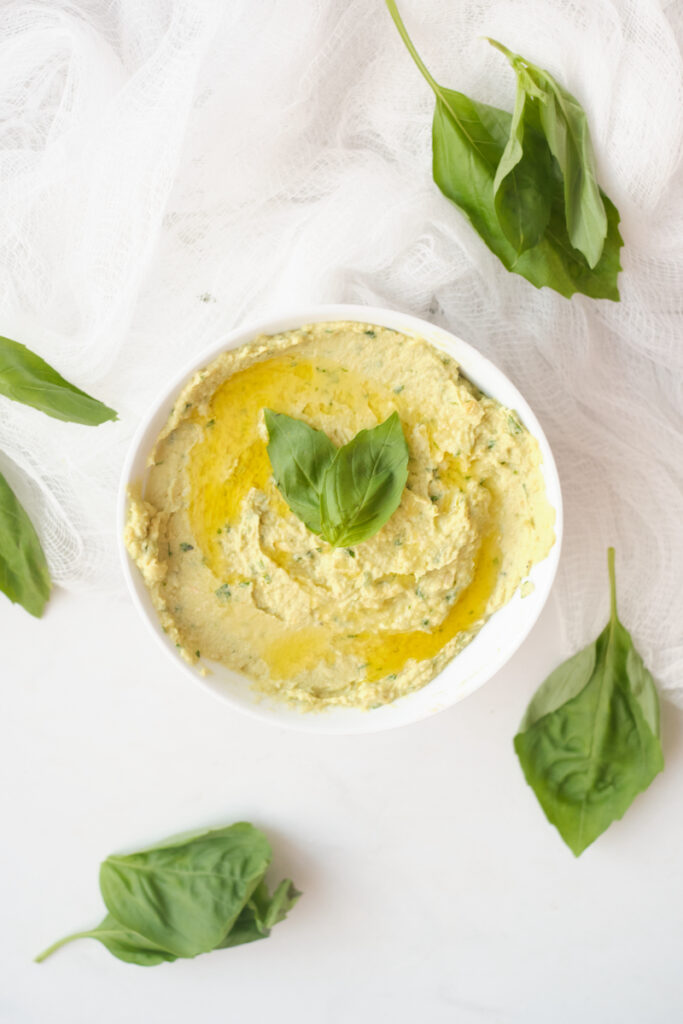 Avocado hummus in a bowl topped with basil leaves and with fresh basil leaves strewn about