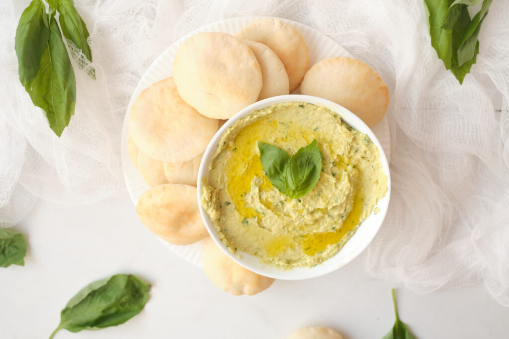 Avocado hummus with basil in a serving bowl surrounded by mini pita rounds