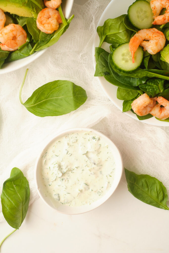 Basil green goddess dressing in a dish surrounded by basil leaves and plates of salad