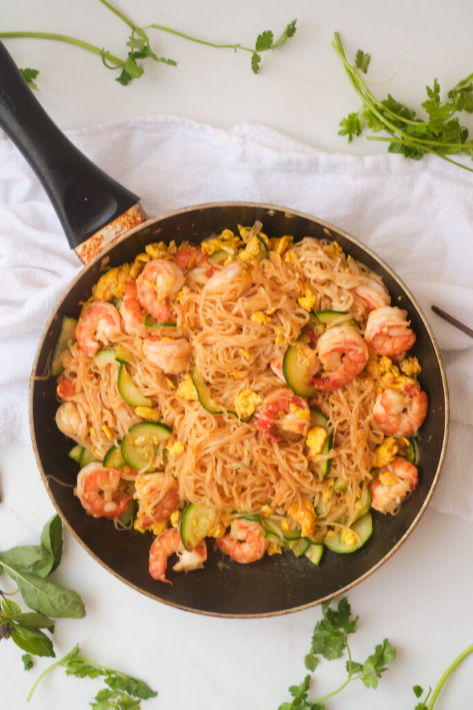 Spicy shrimp noodles in a pan surrounded by loose thai basil