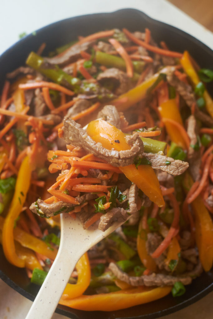 veggie honey ginger beef and basil stirfry in a cast iron pan with a wooden spoon lifting some of it up