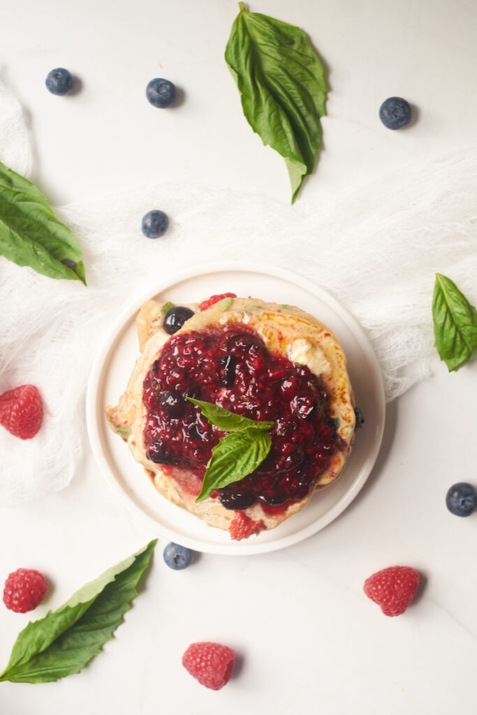 Birds eye view of a stack of lemon basil pancakes topped with butter and basil berry compote and surrounded by berries and basil leaves