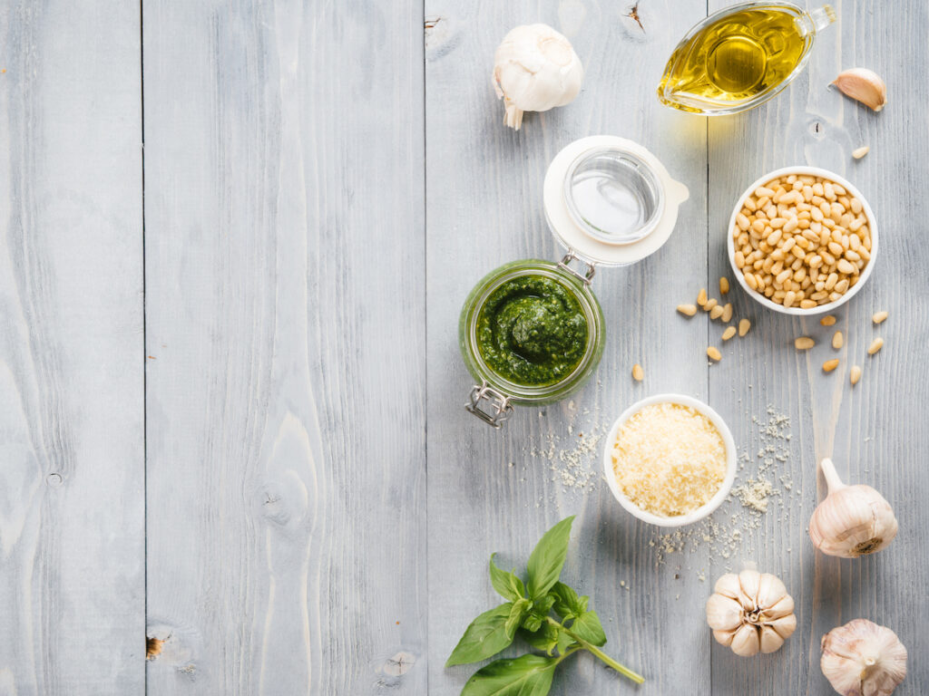 Homemade basil pesto sauce and ingredients on gray wooden background. Classic basil contains pine nuts, which doesn't work for people who need a nut free pesto due to preference or nut allergy. 