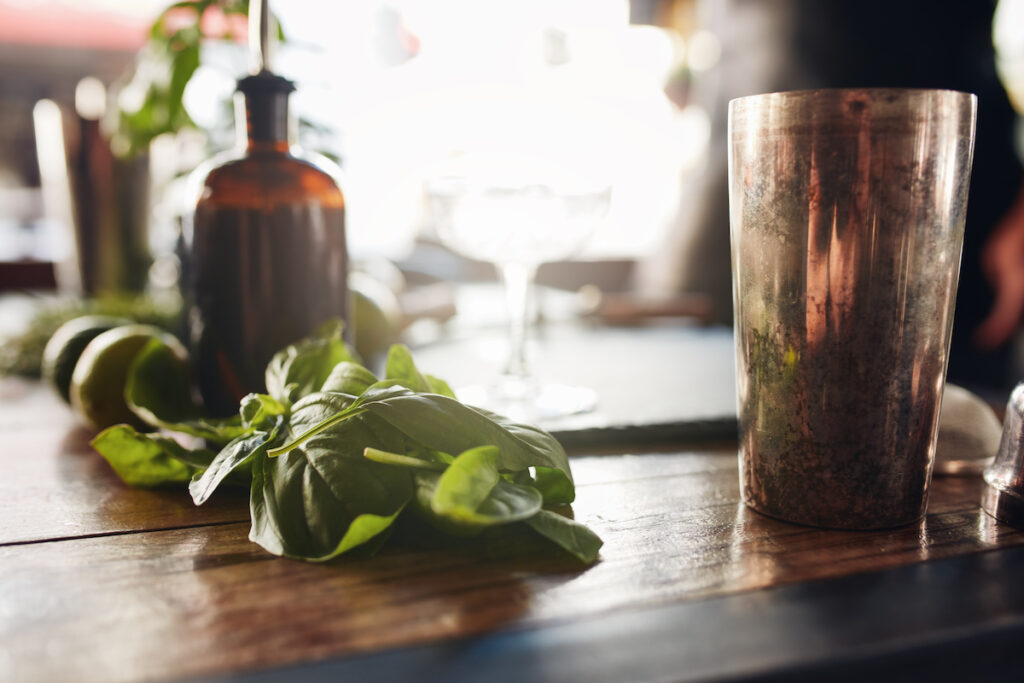 Closeup shot of basil leaves with cocktail shaker and other ingredients on table at bar.