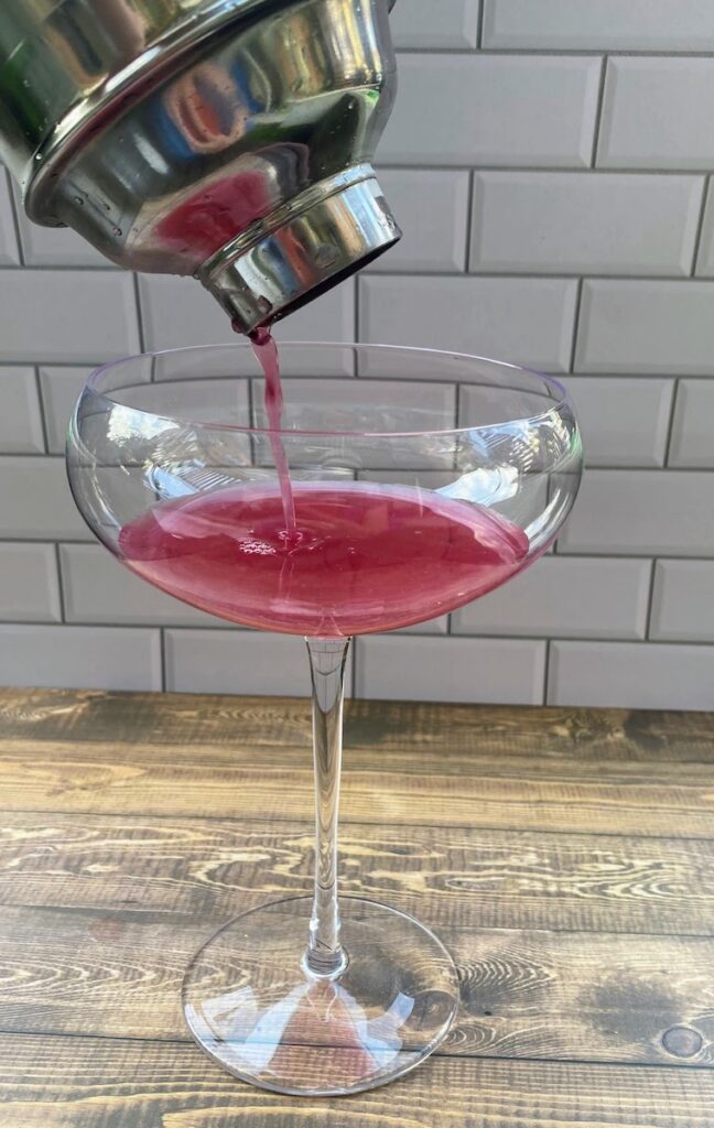 Cocktail shaker pouring purple basil blueberry lemonade with vodka into a cocktail glass