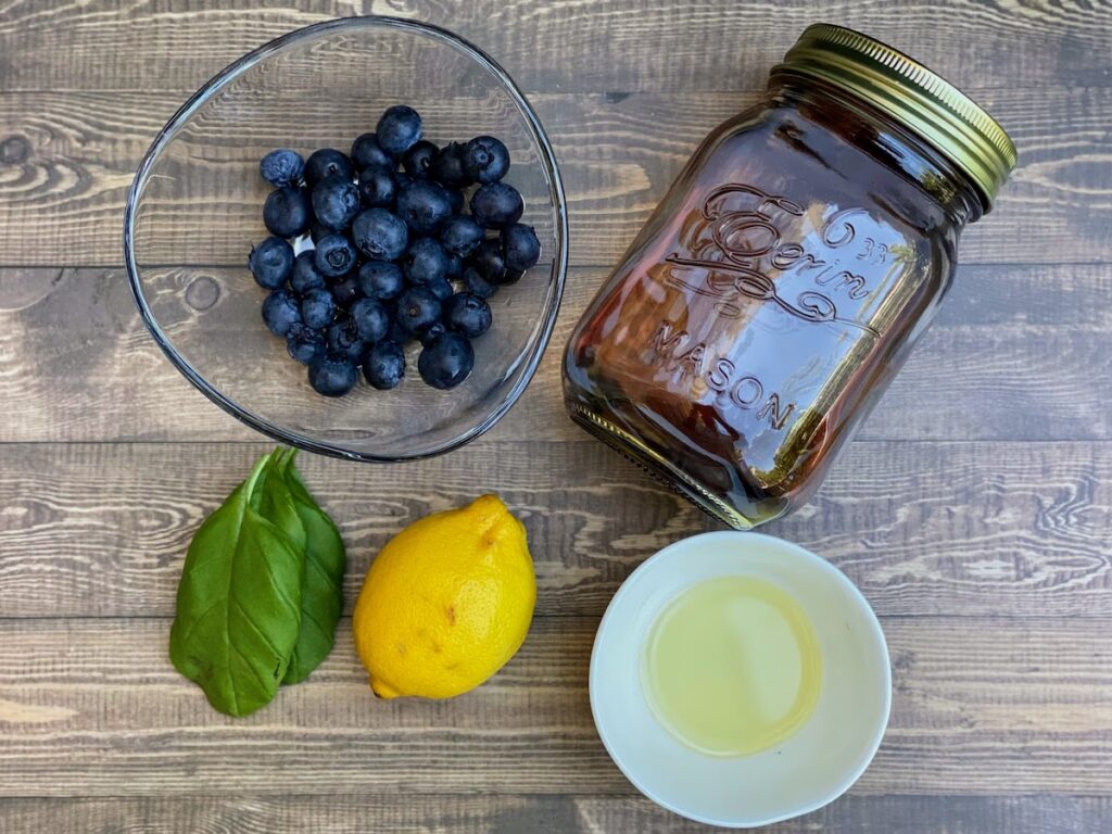 Ingredients to make a basil blueberry vodka lemonade cocktail including infused vodka, simple syrup, lemon, basil and blueberries