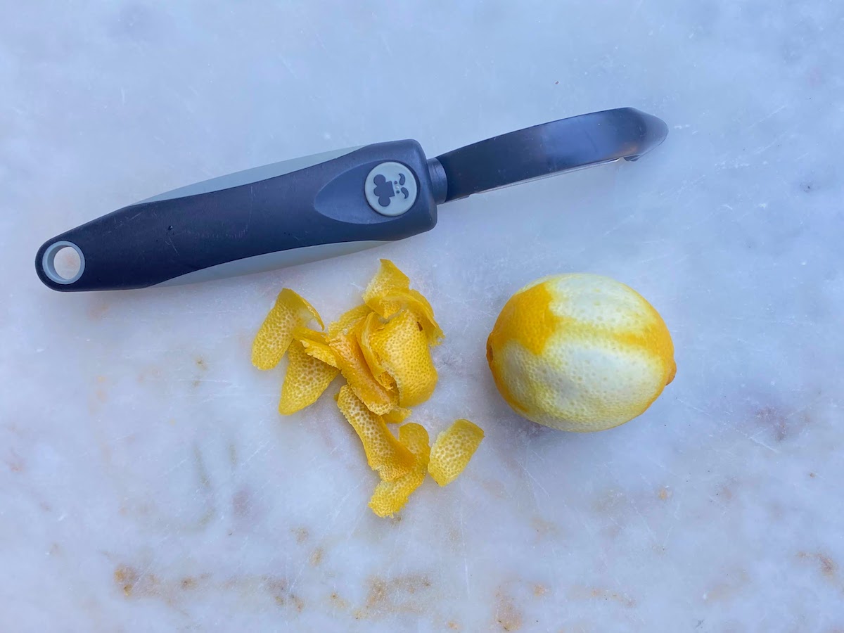 lemon peel, a peeled lemon, and a vegetable peeler on a white marble surface