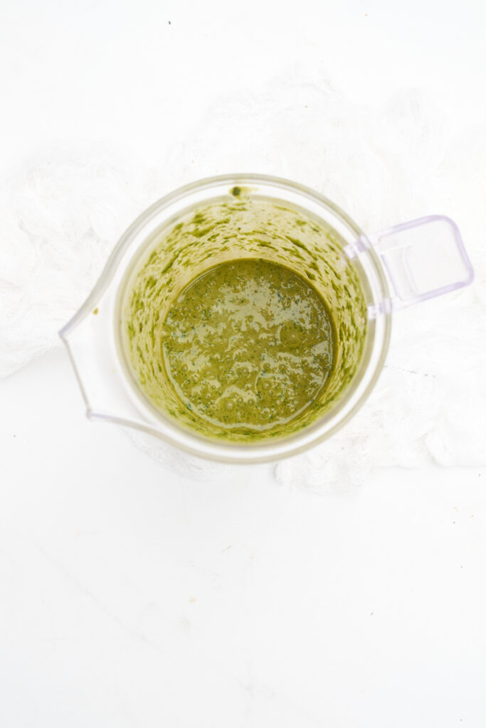 Aerial view of a glass measuring cup sitting on a white surface, filled with fresh basil leaves than have been blended with olive oil into a mixture