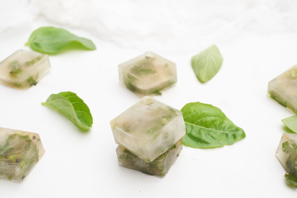Fresh basil leaves and frozen cubes of basil chiffonade frozen in broth are arranged on a white surface. 