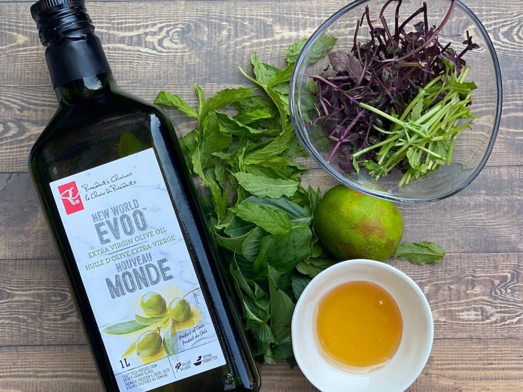 Ingredients for mint and basil stem salad dressing against a wood plank background including EVOO, honey, lime, mint leaves, basil leaves, and raw basil stems or basil stalks