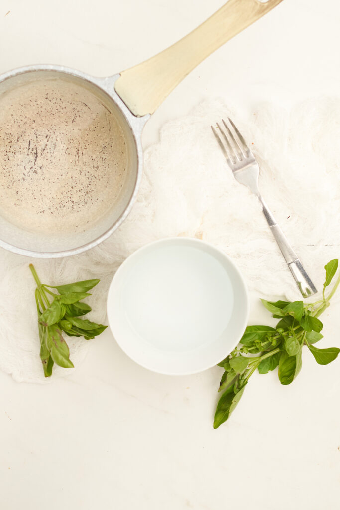 A metal pot, a bowl of cold water, a fork, and fresh basil. Materials needed to blanche fresh basil leaves before freezing