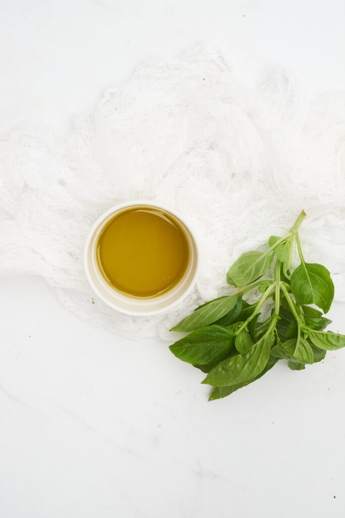 Birds eye view of a stem of fresh basil leaves and a bowl of olive oil on a white counter top