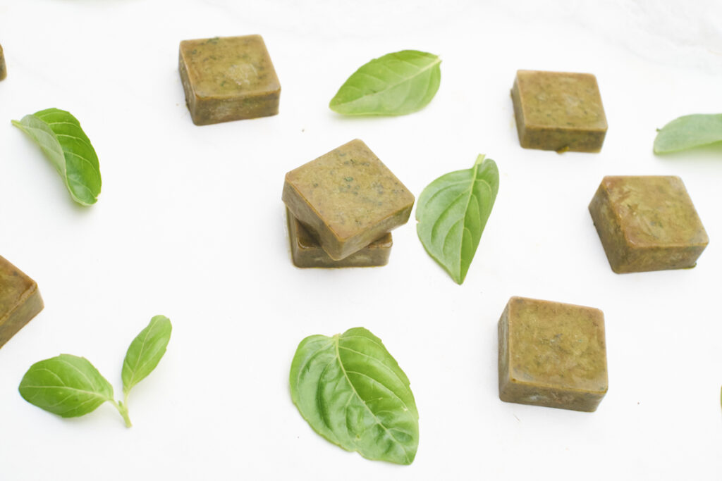Fresh basil leaves and frozen basil and olive oil cubes are arranged artfully on a white surface. 